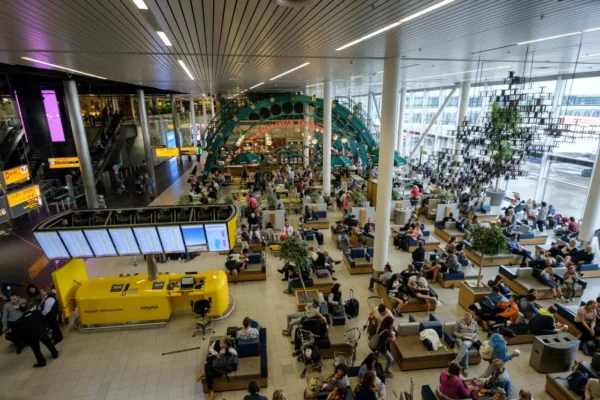People sit and walk through a busy departure hall at Amsterdam Schiphol Airport during daytime.