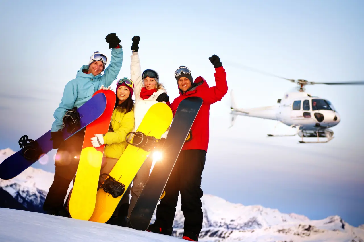 Group of snowboarders celebrating on a snowy mountain ridge with a helicopter landing nearby.