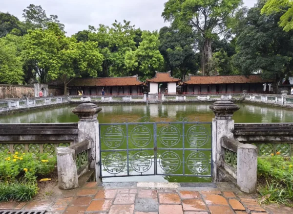 Heavenly Well at the Temple of Literature in Hanoi, with a green reflective pond, stone railings, and traditional tiled-roof buildings surrounded by trees.