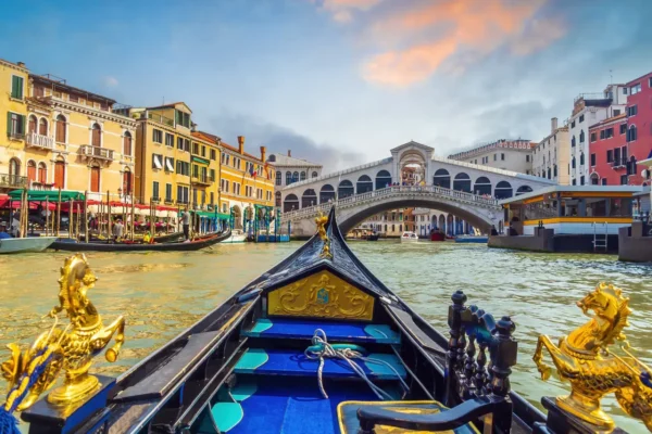 View from a gondola on the Grand Canal toward the Rialto Bridge in Venice, with colorful historic buildings and boats under a bright sky.