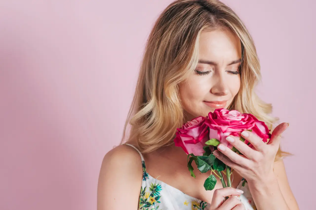 Woman with blonde hair gently smelling a bouquet of pink roses against a soft pastel background.