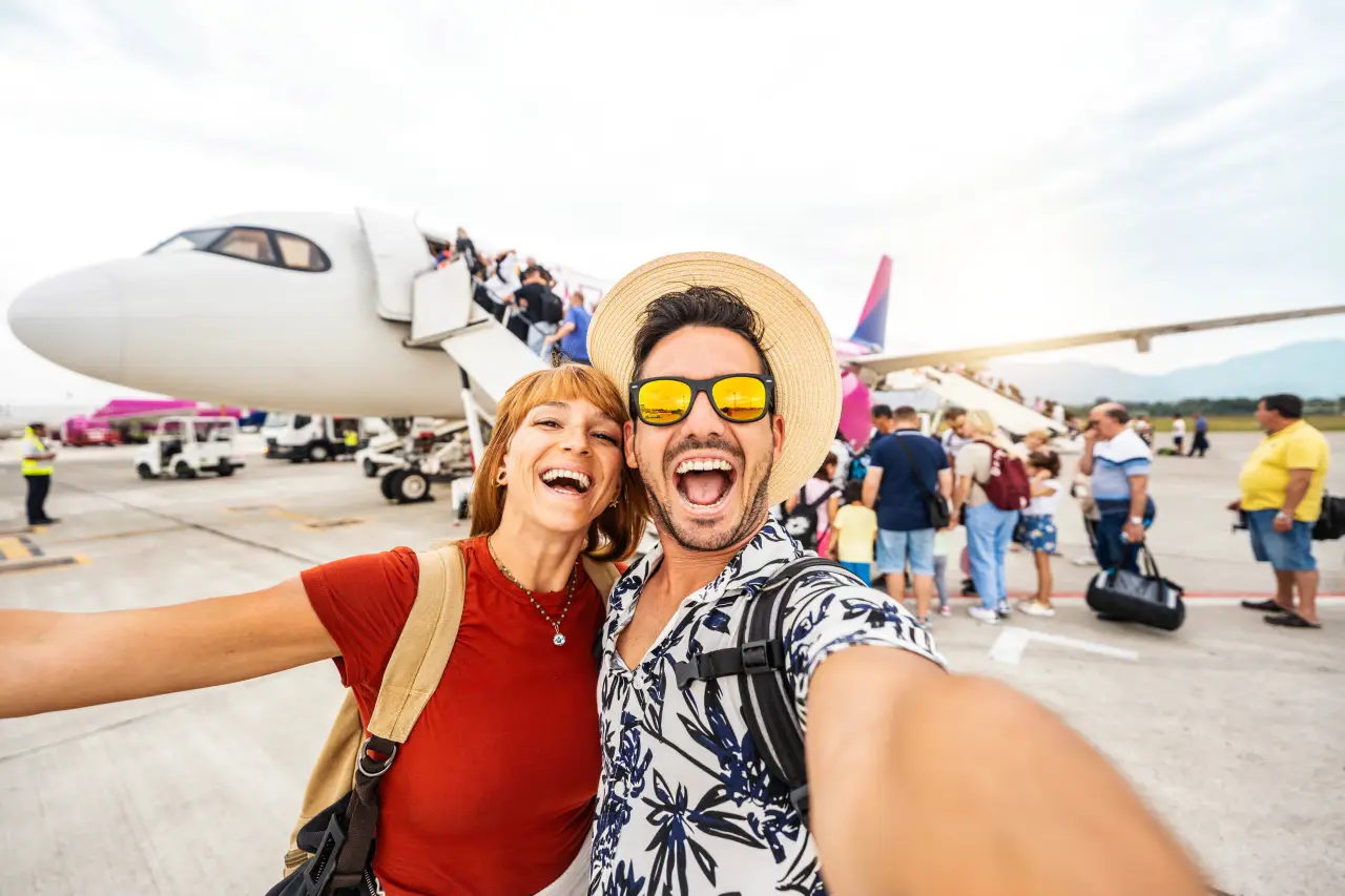 Smiling couple taking a selfie on the airport tarmac while boarding a passenger airplane with other travelers in the background.