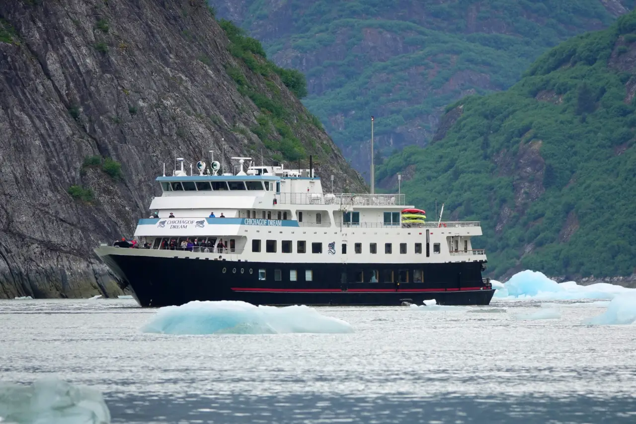 Small expedition cruise ship sailing through icy waters in Southeast Alaska fjord surrounded by steep green mountains and floating ice