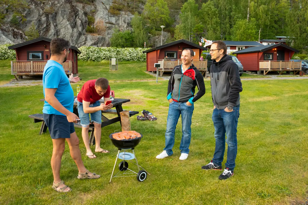 Friends grilling food at a clean, modern campground with wooden cabins and green lawn in spring