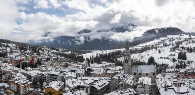 Snow-covered rooftops in Cortina d’Ampezzo with the Basilica Minore dei Santi Filippo e Giacomo rising above the town, framed by the Dolomites and nearby construction cranes.
