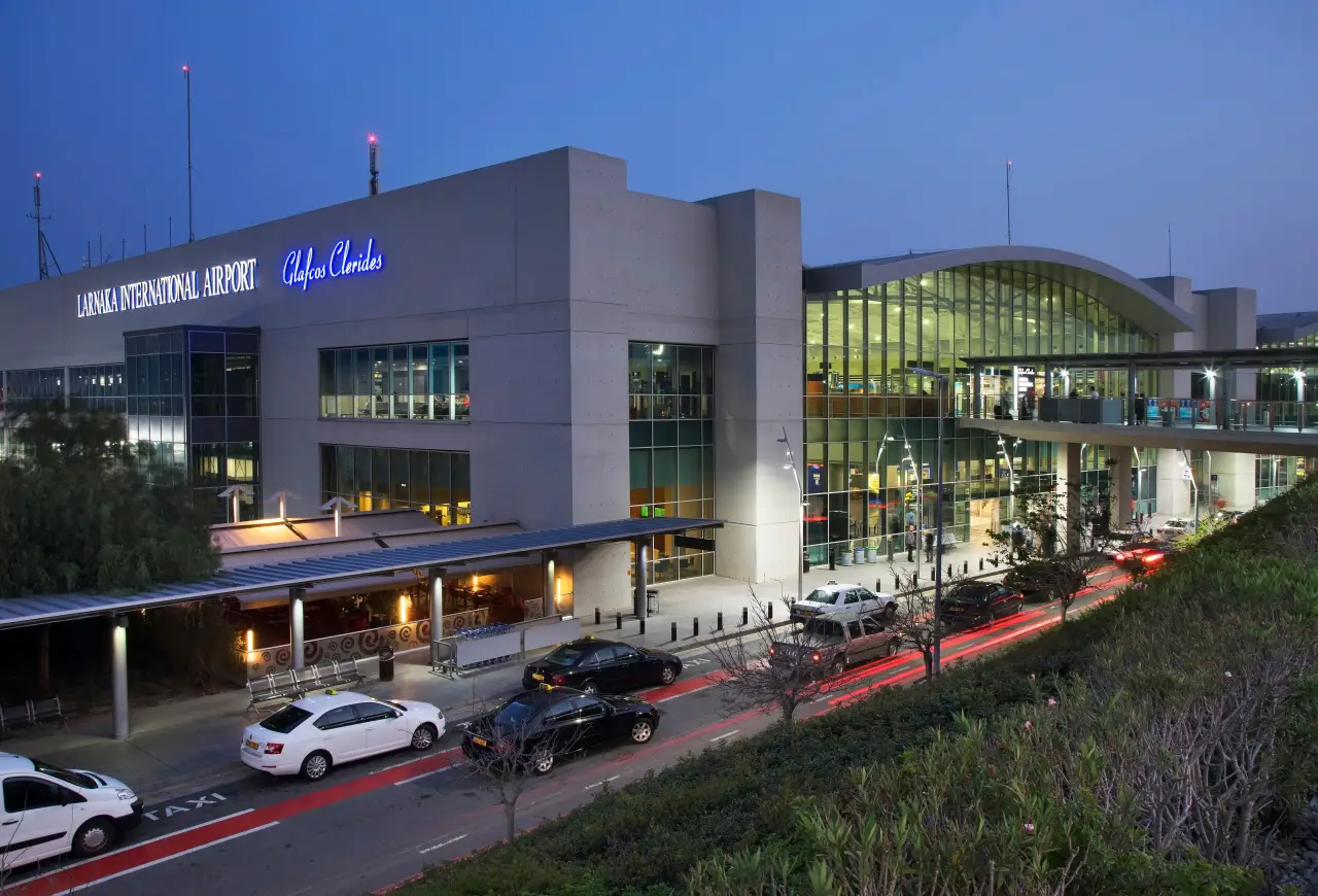 Exterior of Larnaca International Airport terminal in Cyprus at dusk with illuminated glass facade and vehicles outside the arrivals area