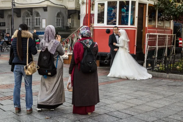 Home 11 Bride and groom posing for wedding photos beside a historic red tram in Istanbul as onlookers take pictures