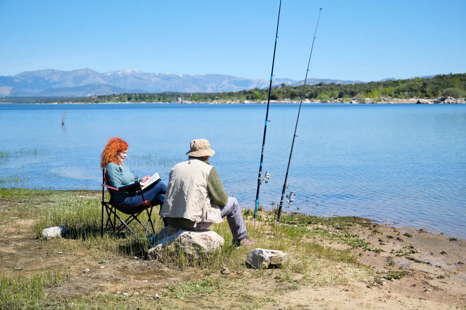 Couple relaxing while fishing on a lakeshore with mountain backdrop on a sunny day