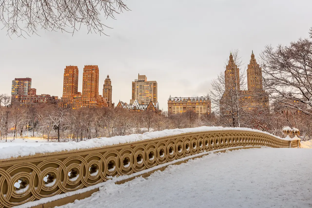 Central Park’s Bow Bridge covered in fresh snow at twilight, with Manhattan’s Upper West Side skyline glowing in the background.
