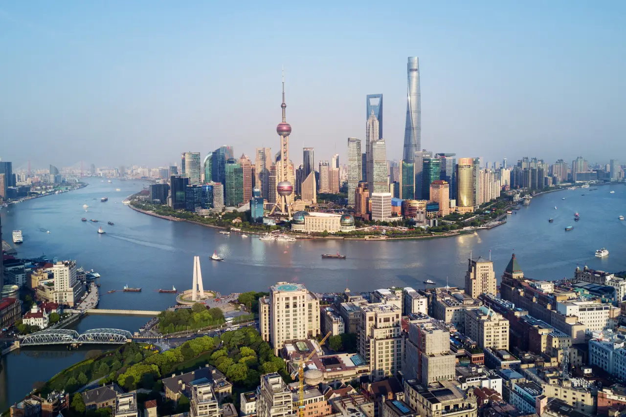 An aerial panoramic view of the Shanghai skyline featuring the Oriental Pearl Tower and Shanghai Tower overlooking the Huangpu River with boats under a clear blue sky.