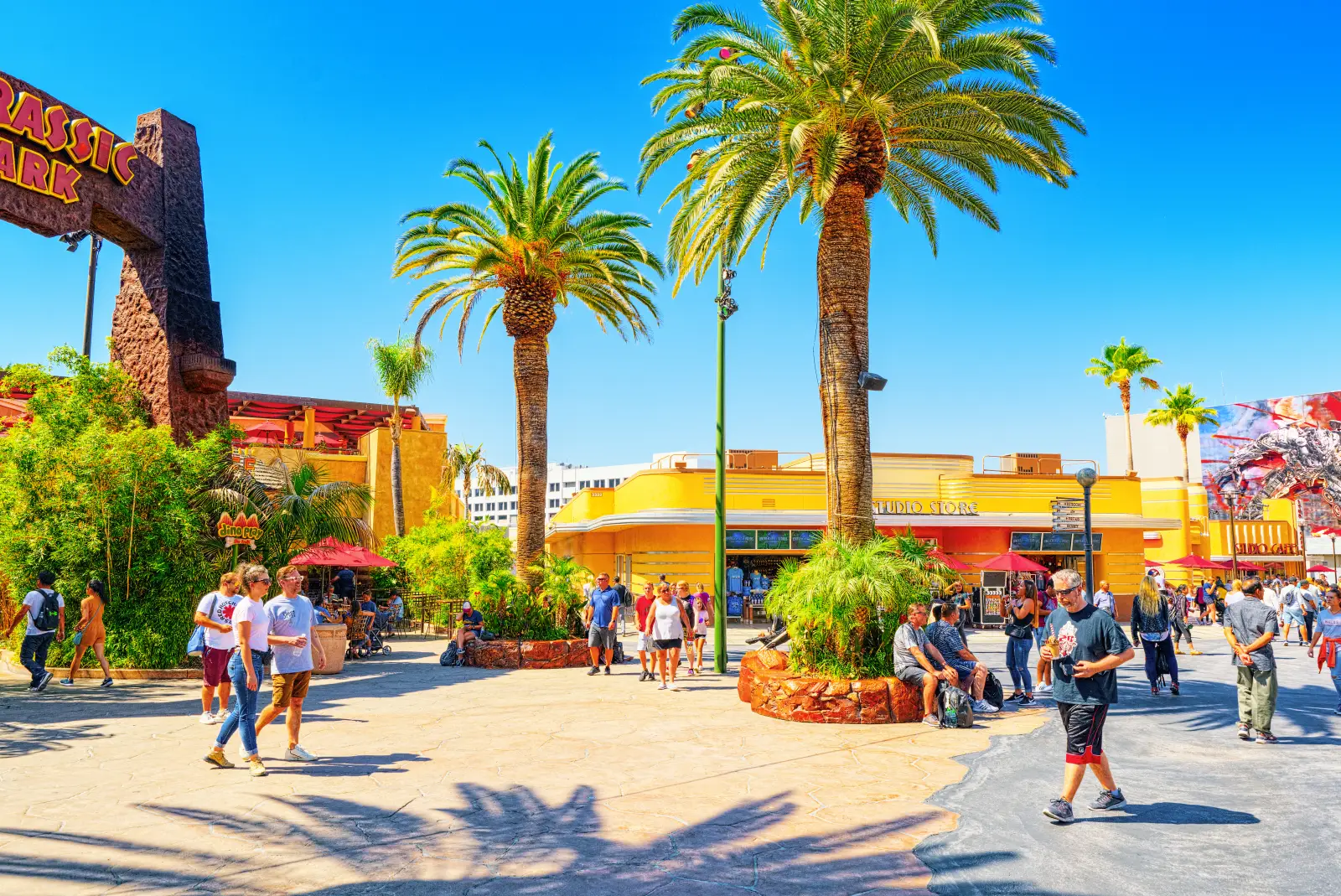 Crowds walking through Universal Studios Hollywood theme park plaza with palm trees and Jurassic Park entrance on a sunny day