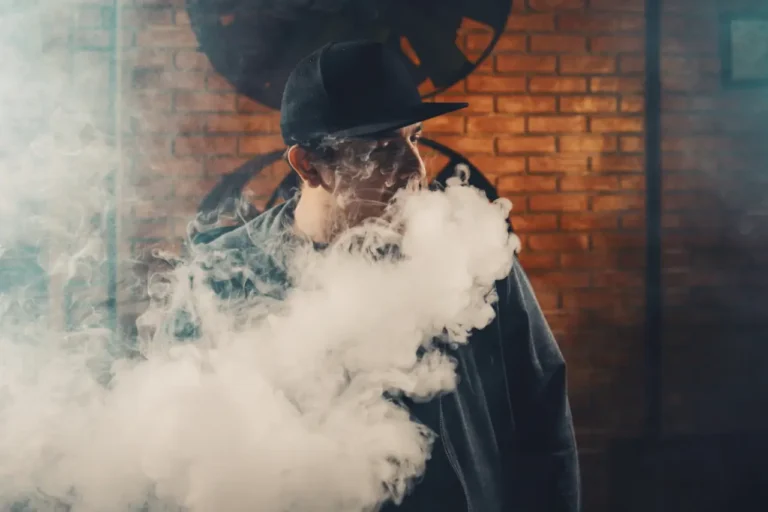 Man exhaling thick vape cloud indoors against brick wall background