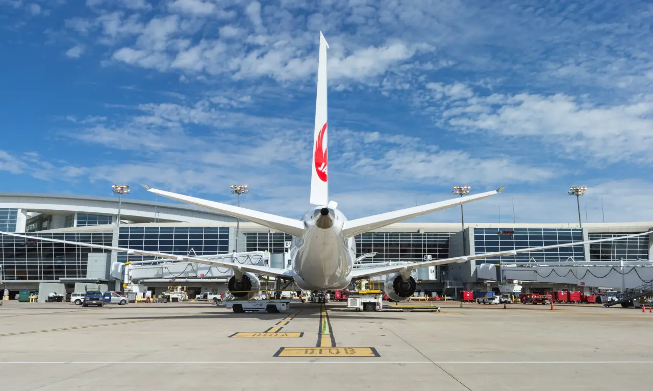Rear view of a Japan Airlines aircraft parked at a gate at Dallas Fort Worth International Airport with terminal building and blue sky.