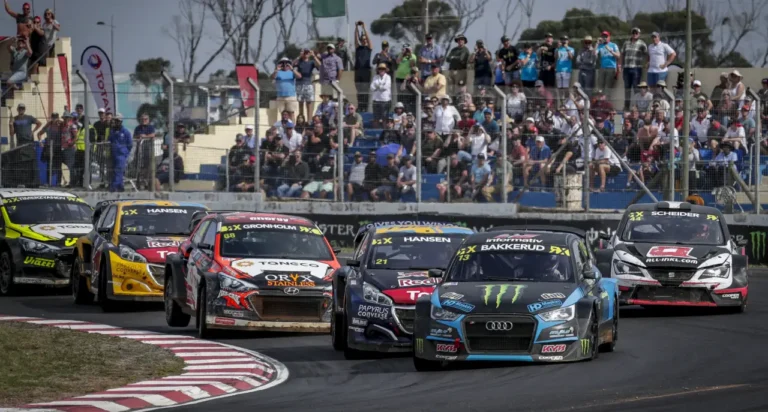 Rallycross cars competing in a close wheel-to-wheel race on a circuit track with spectators watching from grandstands.