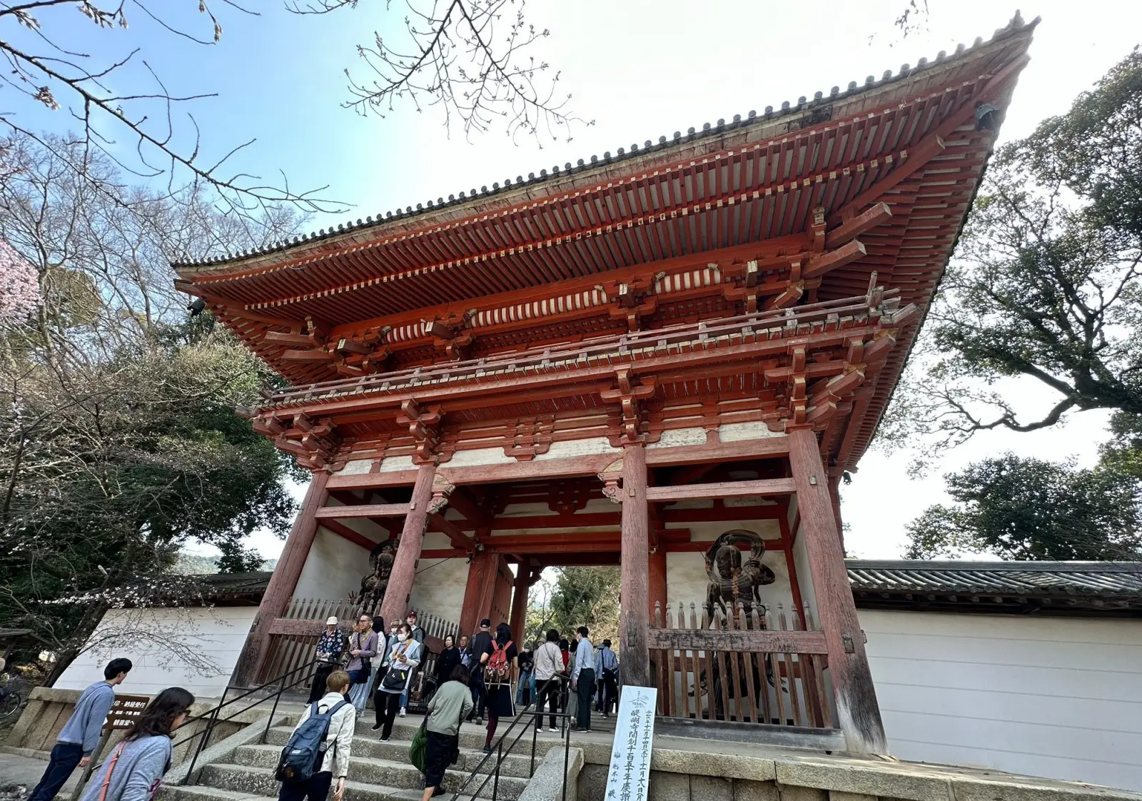 Visitors entering a traditional Japanese temple gate with wooden architecture and cherry blossoms in the background