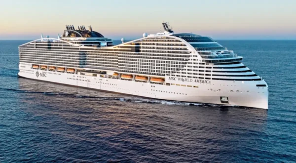 Home 11 MSC World America cruise ship sailing at sunset on open sea, showcasing the modern flagship vessel of MSC Cruises with multiple decks and balcony cabins.