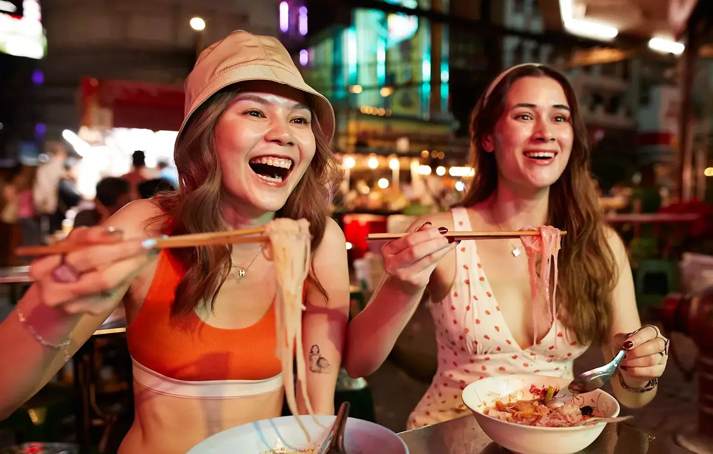 Two travelers enjoying street food with chopsticks at a lively night market in Bangkok, showcasing Asia’s vibrant street food culture