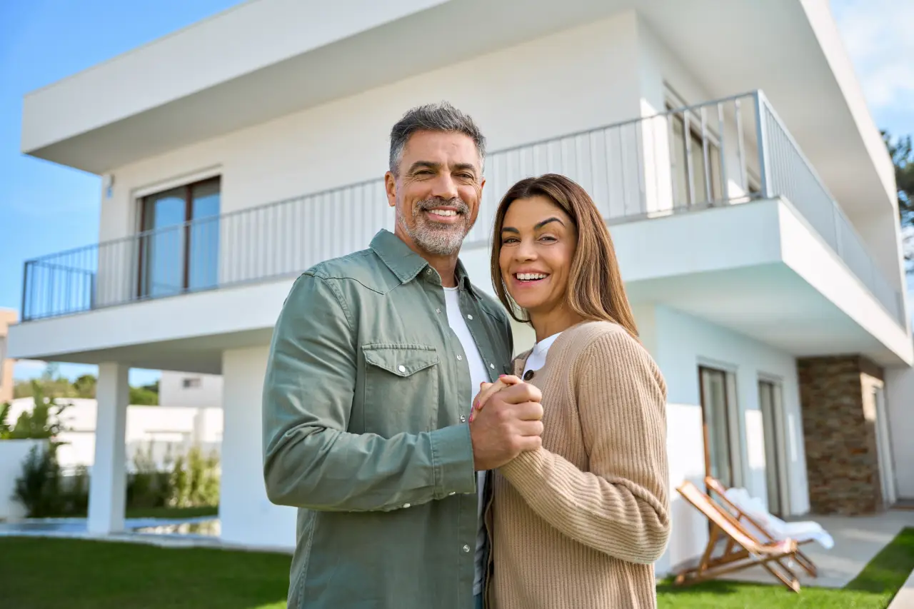 Smiling couple standing in front of a modern white house with a balcony and garden