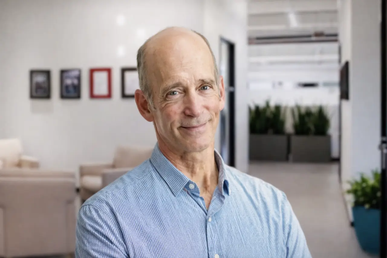 Portrait of Dr. Mercola standing indoors in a modern office setting with neutral decor and plants in the background