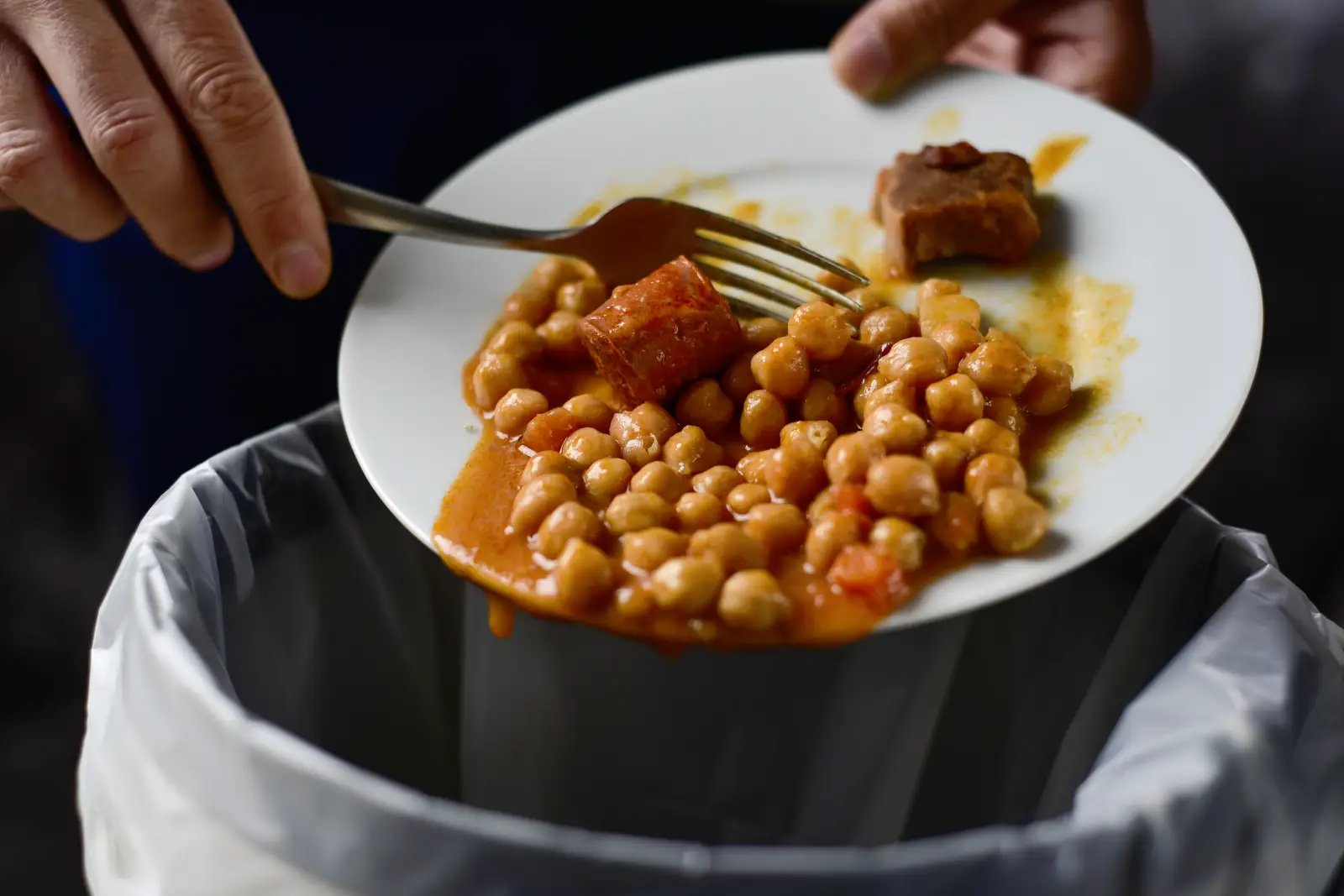 Close-up of a plate of leftover chickpea stew with meat being scraped into a trash bin, highlighting food waste