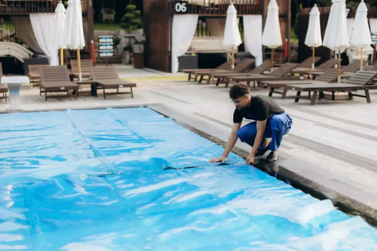 Technology 6 A pool maintenance worker crouching at the edge of an outdoor hotel swimming pool, pulling a blue thermal cover over the water, with lounge chairs and white umbrellas in the background.