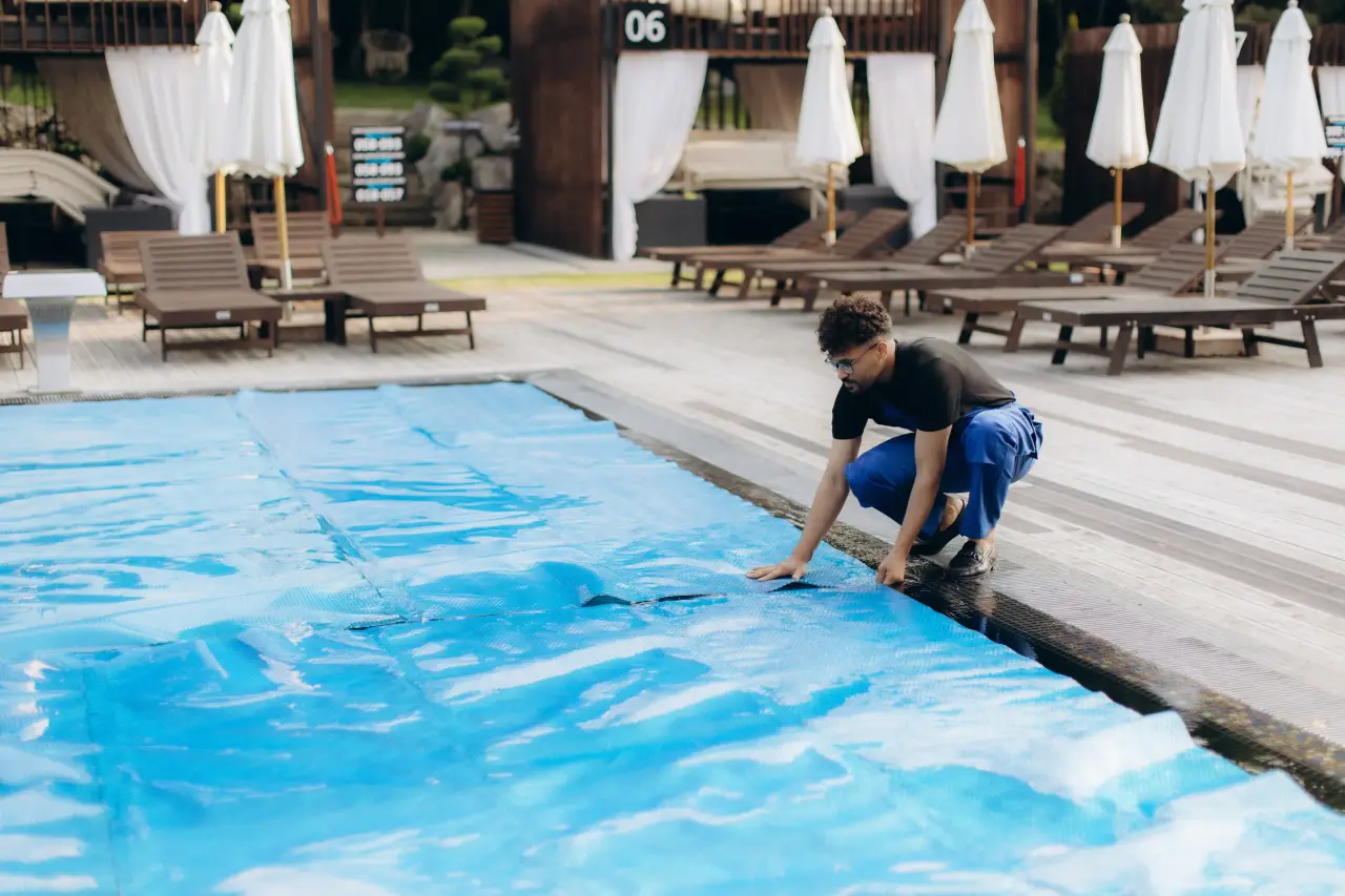 A pool maintenance worker crouching at the edge of an outdoor hotel swimming pool, pulling a blue thermal cover over the water, with lounge chairs and white umbrellas in the background.