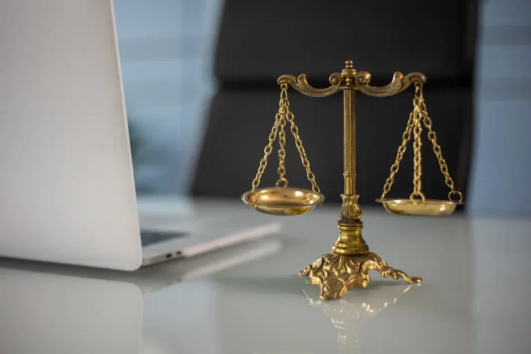 Other News 19 Close-up of gold balance scales on a desk beside a laptop, symbolizing legal compliance and business law in a modern office setting