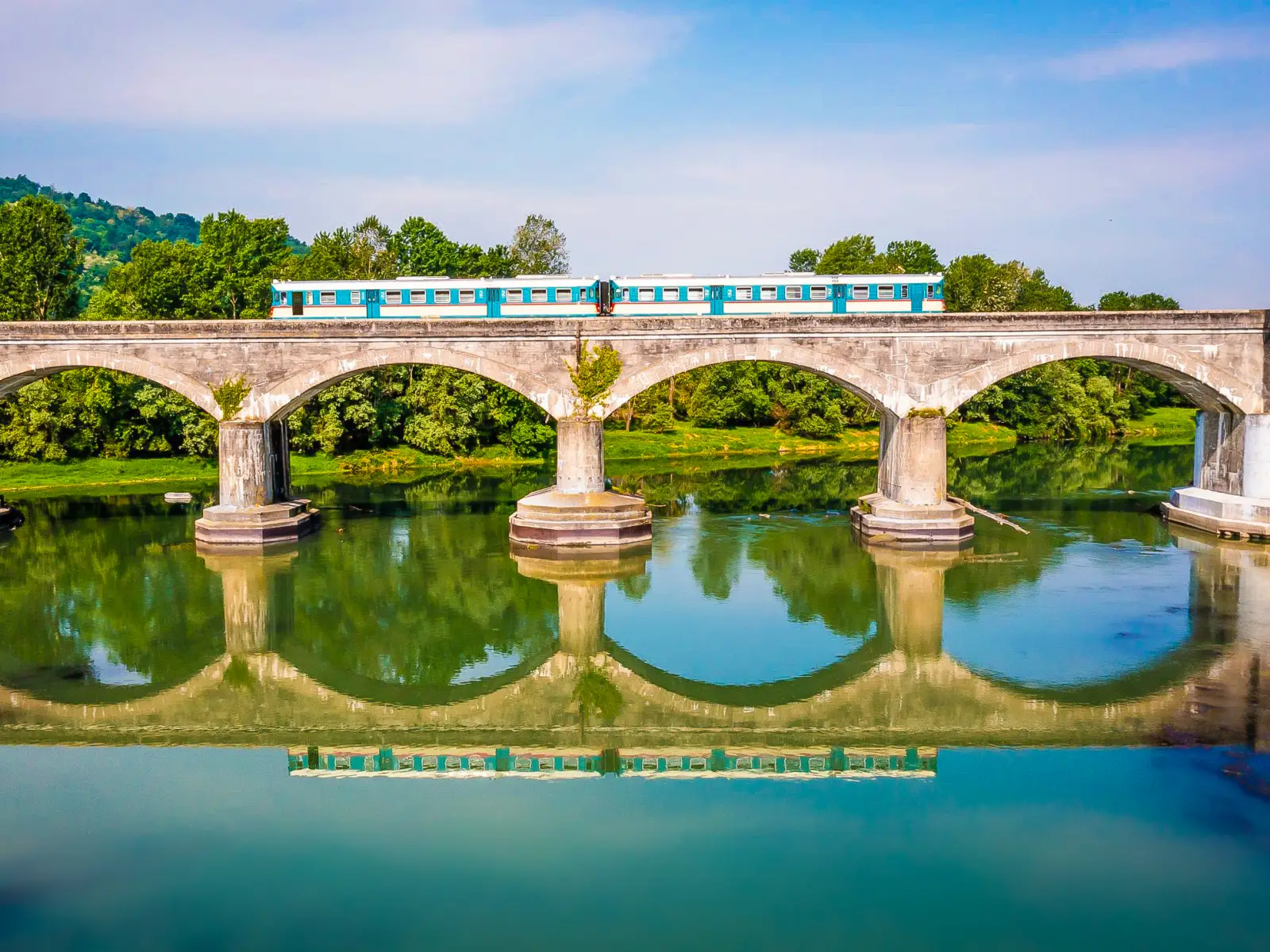 Vintage turquoise train crossing a stone railway bridge over a calm river in Lombardy, Italy, reflecting scenic slow travel experience