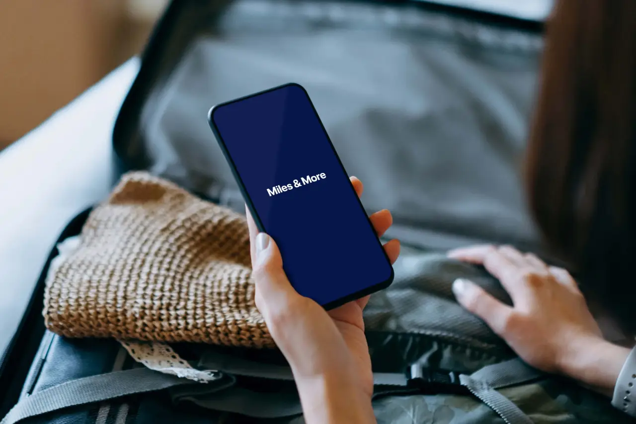 Passenger holding smartphone displaying Miles and More loyalty program app while packing suitcase for travel