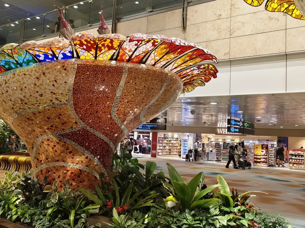 Colorful mosaic sculpture installation inside Singapore Changi Airport terminal with shops and travelers in the background