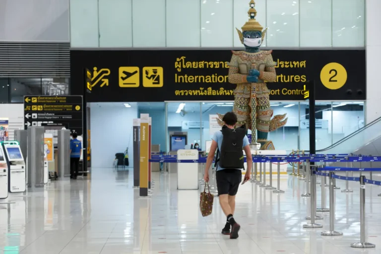Tours 17 Traveler walking through a quiet international departures hall at a Thailand airport with a masked guardian statue and signage overhead