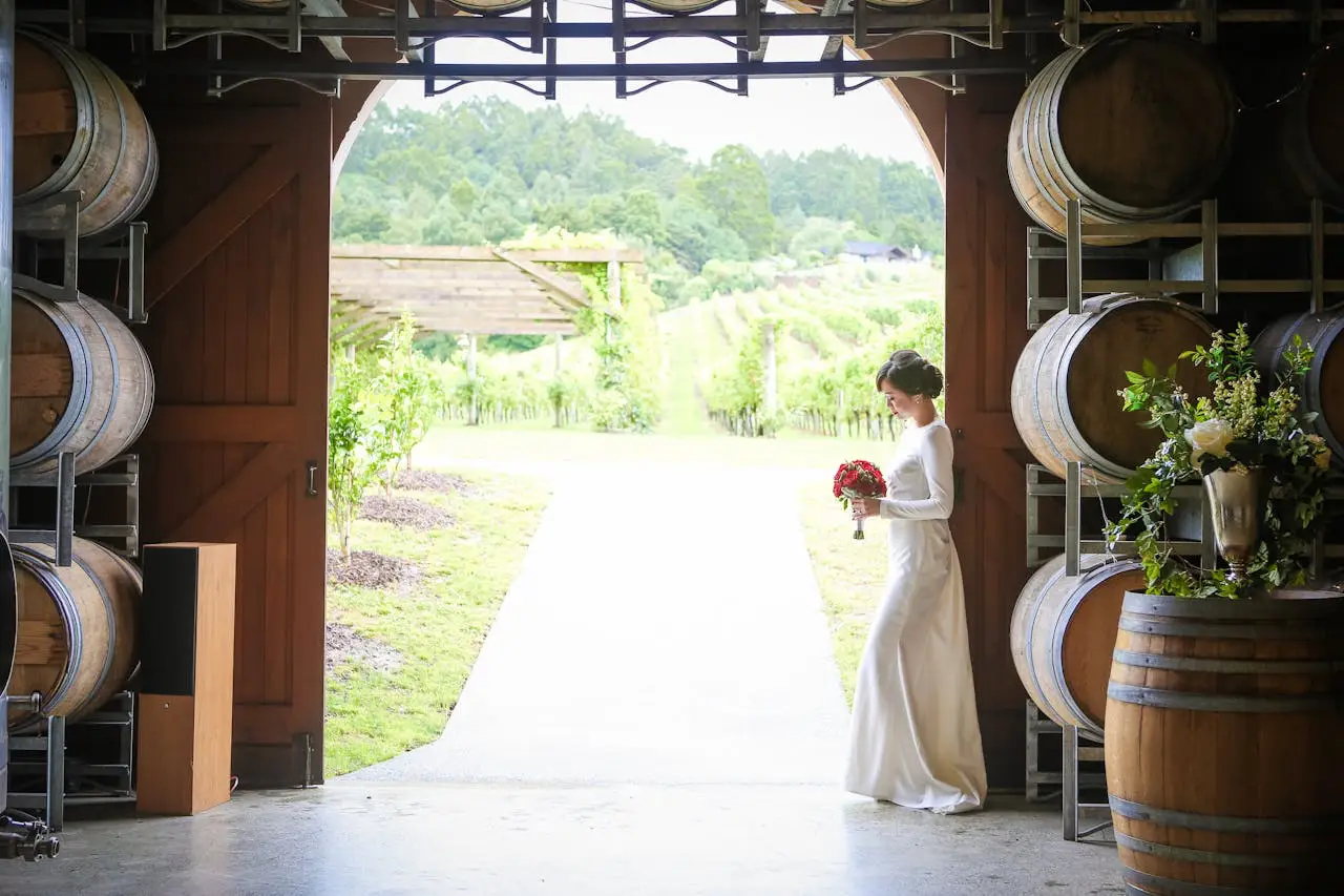 Bride in a white dress holding a bouquet inside a winery, with vineyard views through open barn doors