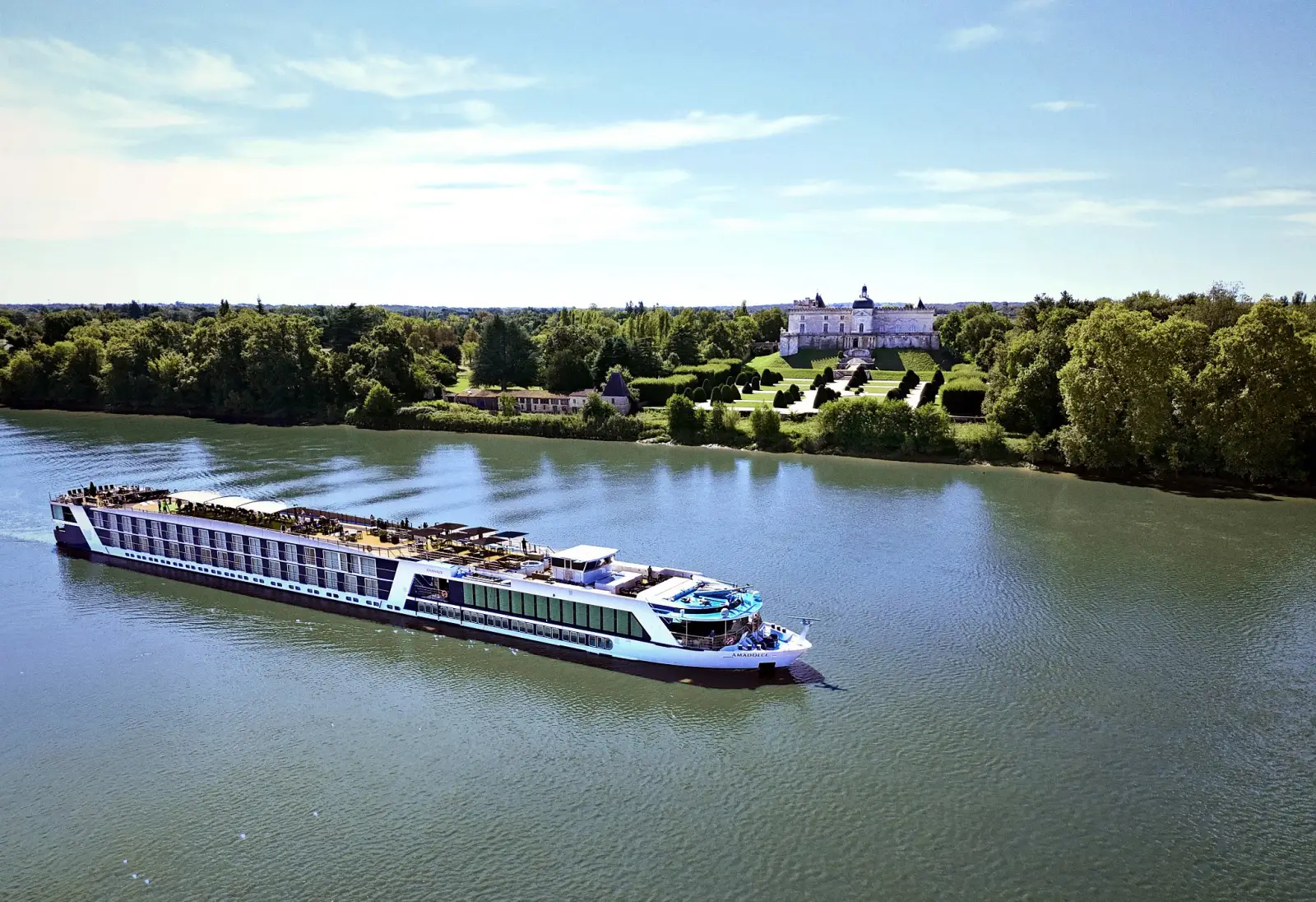 Luxury AmaWaterways river cruise ship sailing past a historic château along a scenic European river under clear blue skies