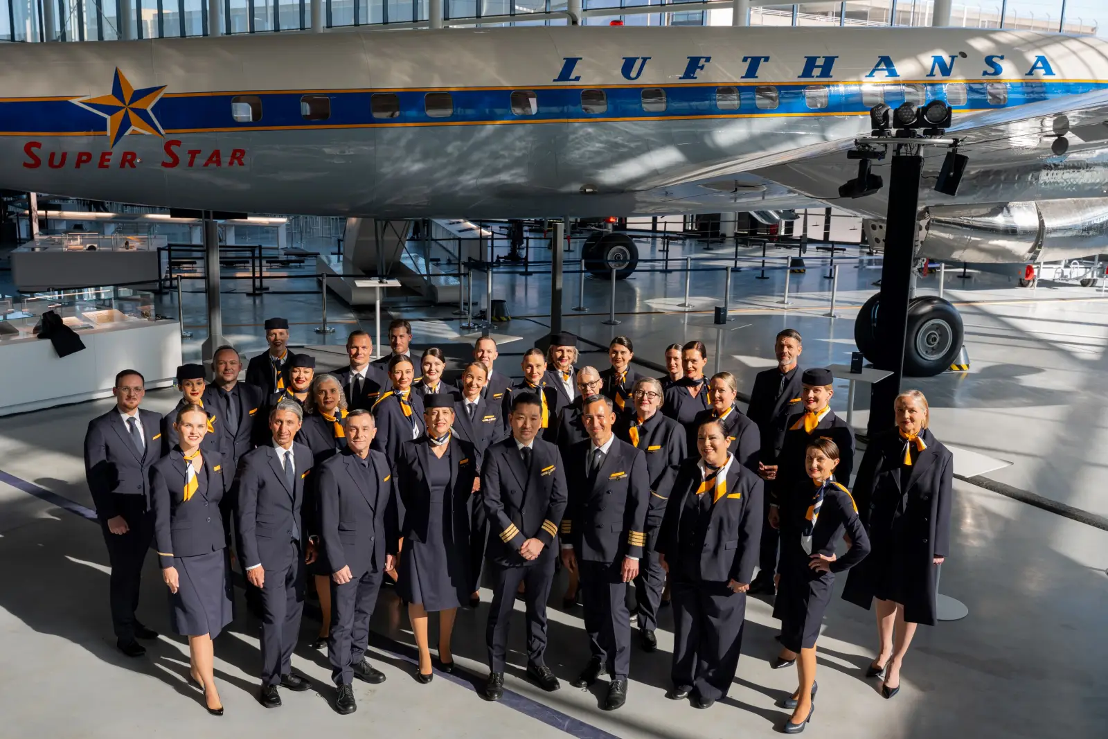 Lufthansa crew members pose in new uniforms inside an aviation hall beside a historic Lufthansa Super Star aircraft.