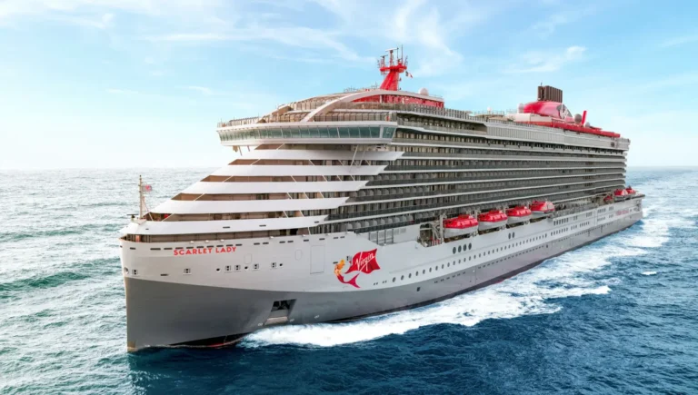 The Virgin Voyages cruise ship Scarlet Lady sailing at sea, viewed from a low aerial angle, showing its white hull, red Virgin logo, and multiple passenger decks under a clear blue sky