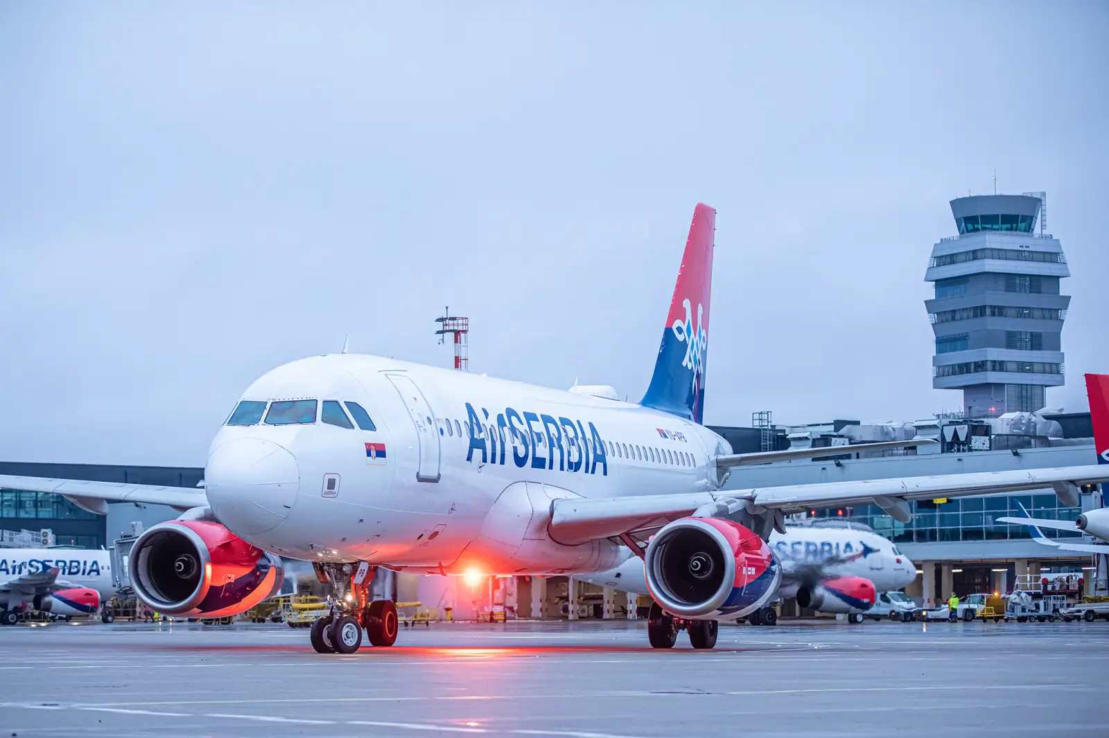Air Serbia Airbus A319 aircraft taxiing on wet airport runway at dusk with terminal and control tower in background