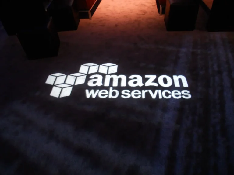 The Amazon Web Services logo projected in white light onto a dark carpeted floor at a corporate event, with warmly lit seating visible in the background