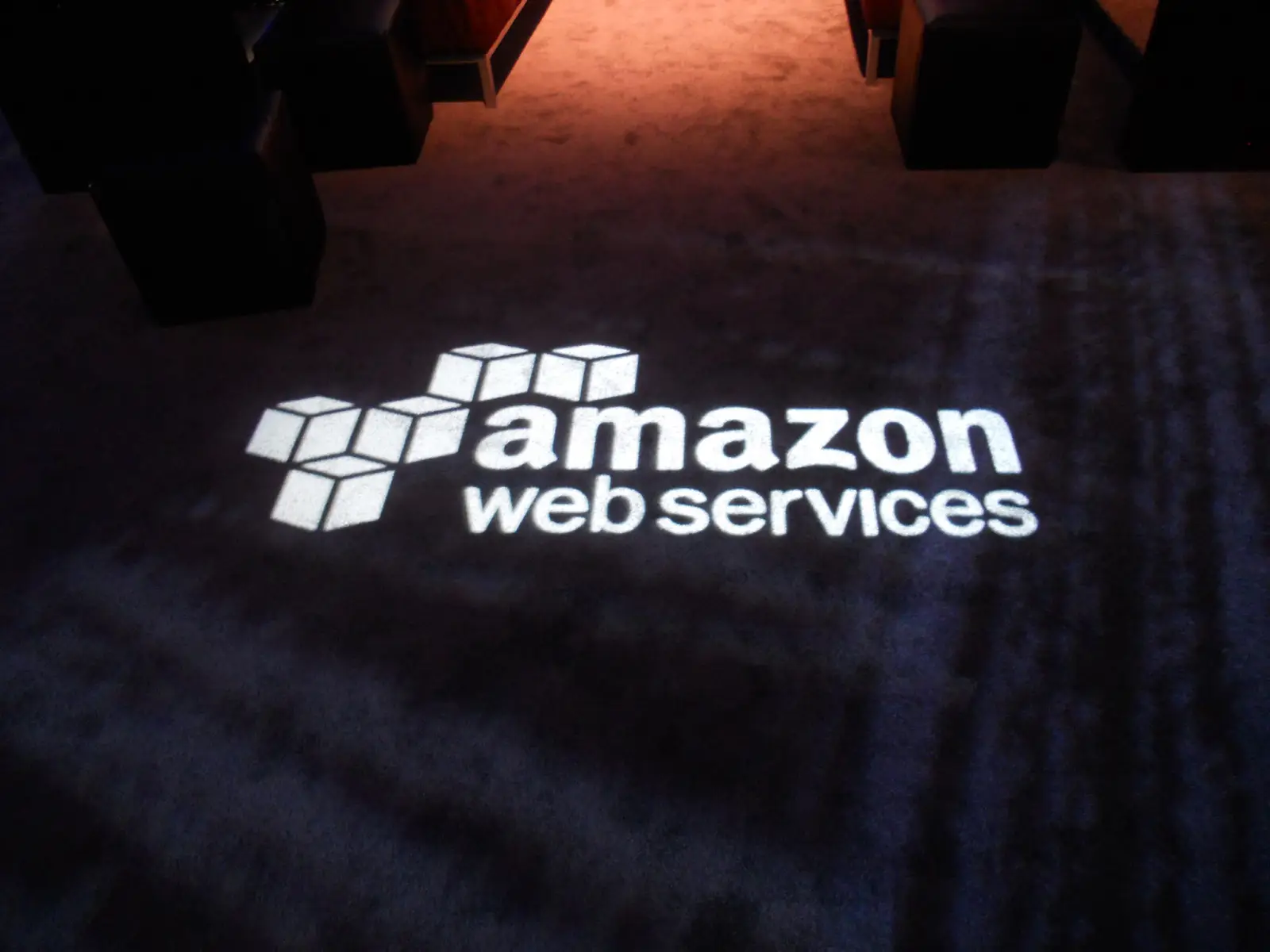 The Amazon Web Services logo projected in white light onto a dark carpeted floor at a corporate event, with warmly lit seating visible in the background