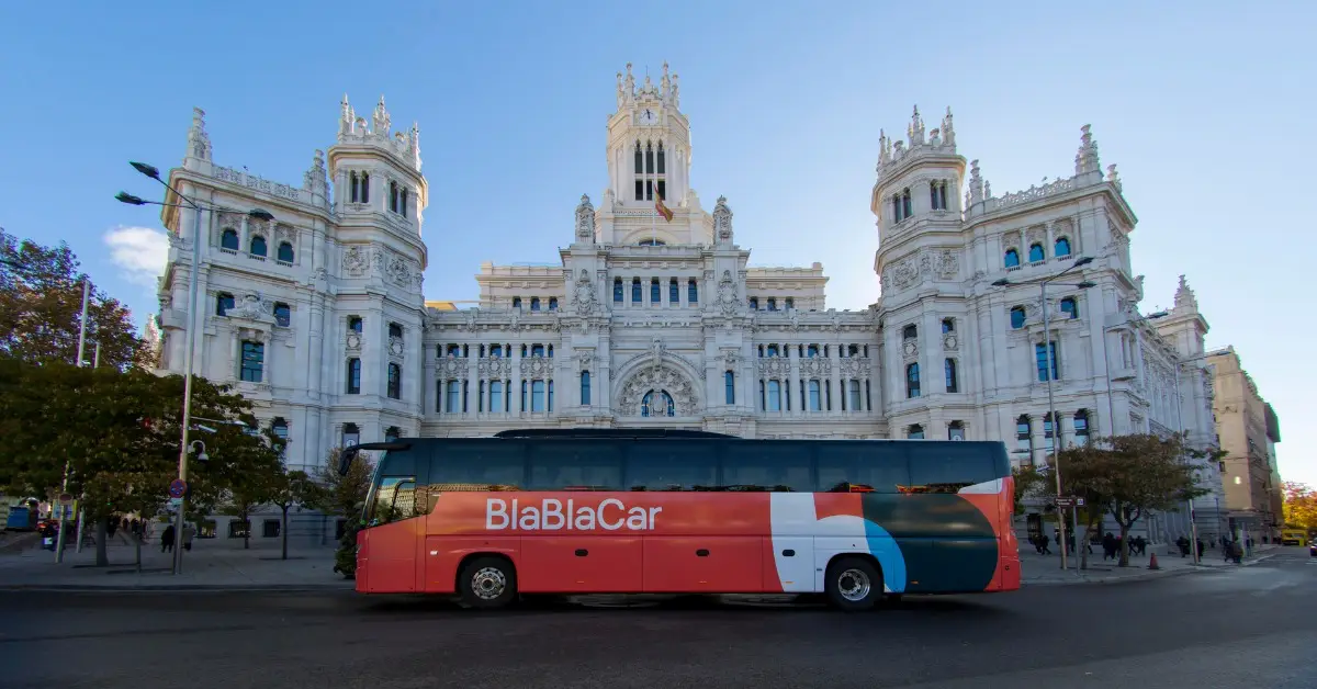 A BlaBlaCar-branded coach in orange, white and teal livery parked in front of the ornate white facade of the Palacio de Cibeles in Madrid, Spain, under a clear blue sky