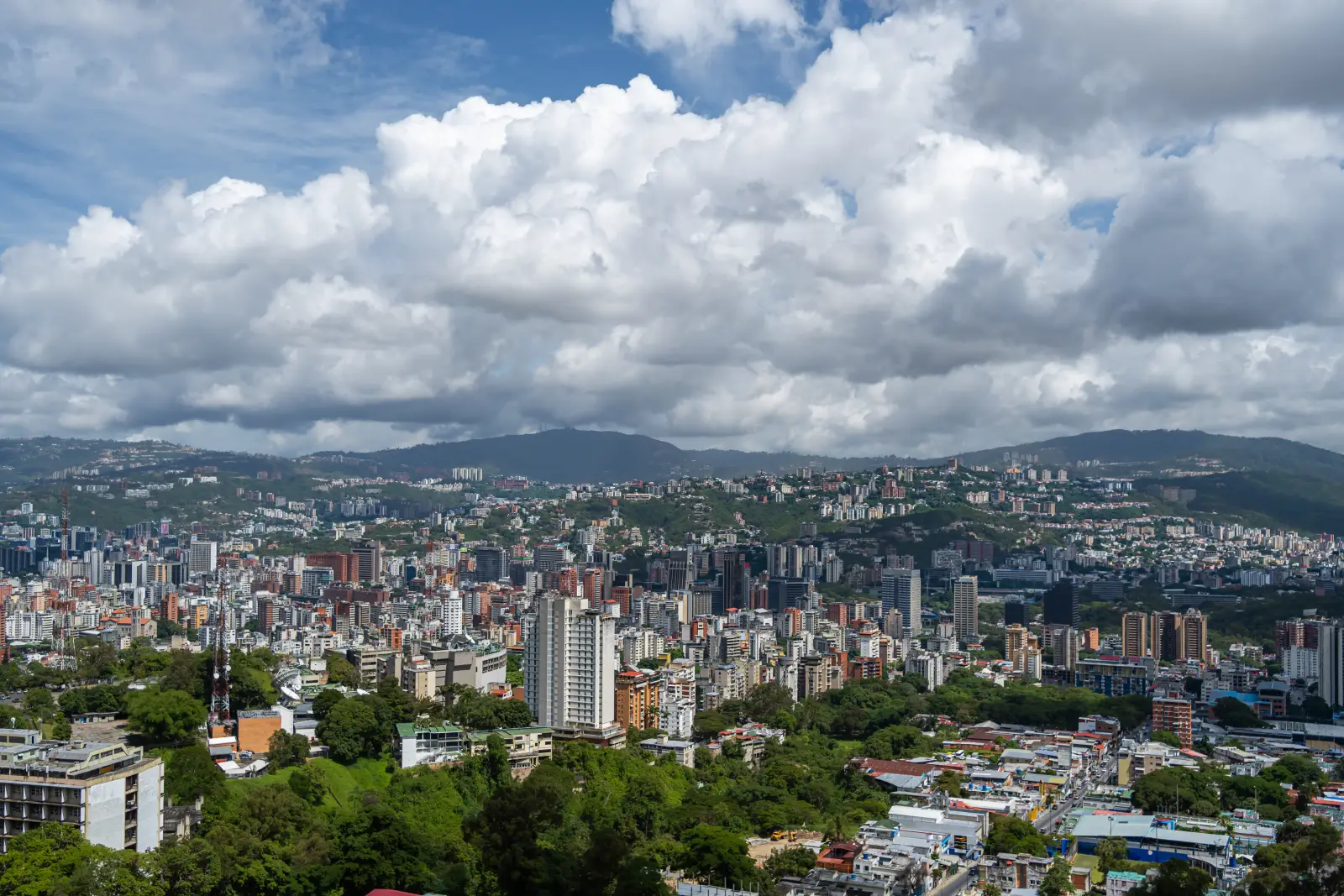 Panoramic aerial view of Caracas, Venezuela, showing a dense urban skyline of residential and commercial buildings surrounded by green hills and mountains under a partly cloudy sky