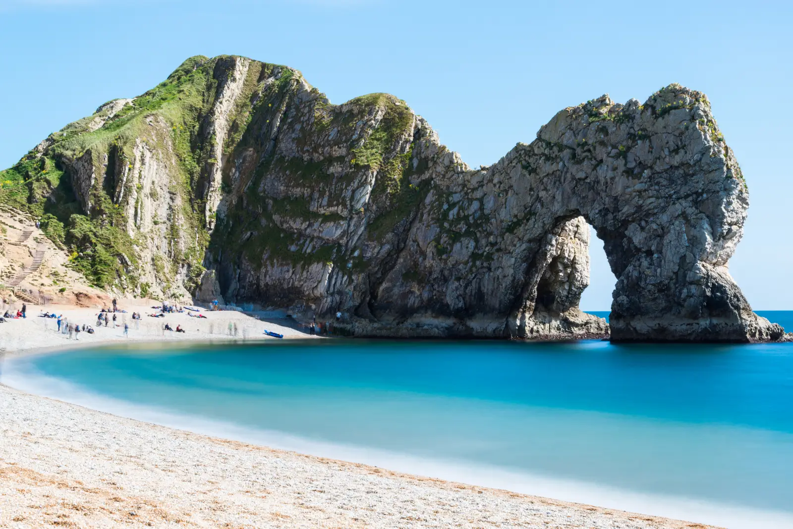 A wide-angle view of Durdle Door, the natural limestone arch on the Jurassic Coast in Dorset, England, with turquoise water, a pebble beach and visitors in the foreground under a clear blue sky