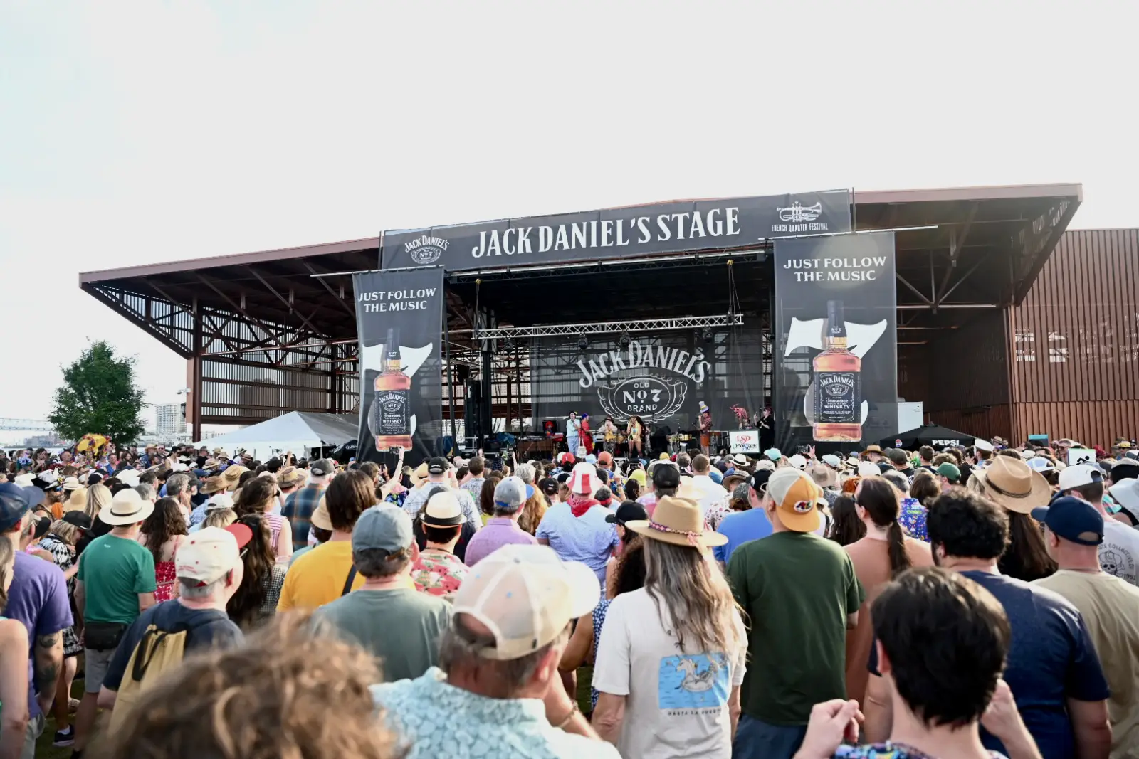 A large crowd of festivalgoers in hats and summer clothing faces the Jack Daniel's Stage at the 2026 French Quarter Festival, with performers visible on stage beneath a black canopy bearing Jack Daniel's Old No. 7 branding and the tagline "Just Follow the Music," set against a hazy sky at Goldring Woldenberg Riverfront Park in New Orleans.