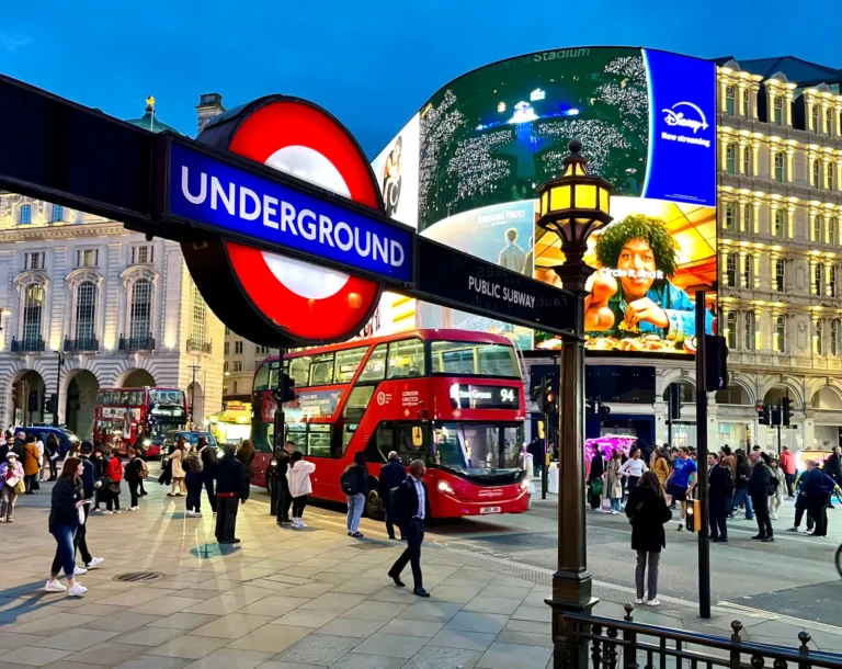 London Underground sign at Piccadilly Circus with red double-decker bus and crowds in central London at dusk