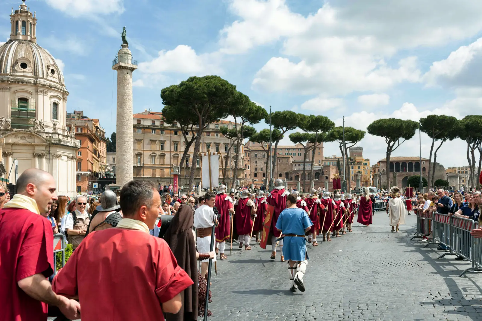 Rome Is Turning 2,779 Today, And The Birthday Celebrations Are Spectacular 3 A long column of costumed Roman reenactors in burgundy cloaks and armour marches along Via dei Fori Imperiali during the Natale di Roma parade, with crowds of spectators behind barriers, Trajan's Column and the Church of the Most Holy Name of Mary visible in the background