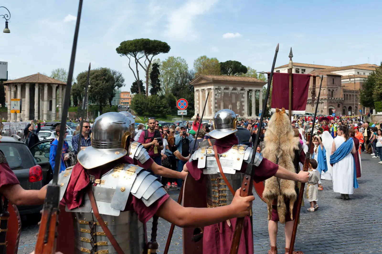 Costumed Roman legionary reenactors in full armour and burgundy tunics carrying spears and shields march through a cobblestone street in central Rome during the Natale di Roma parade, with crowds of spectators and ancient Roman temples visible in the background