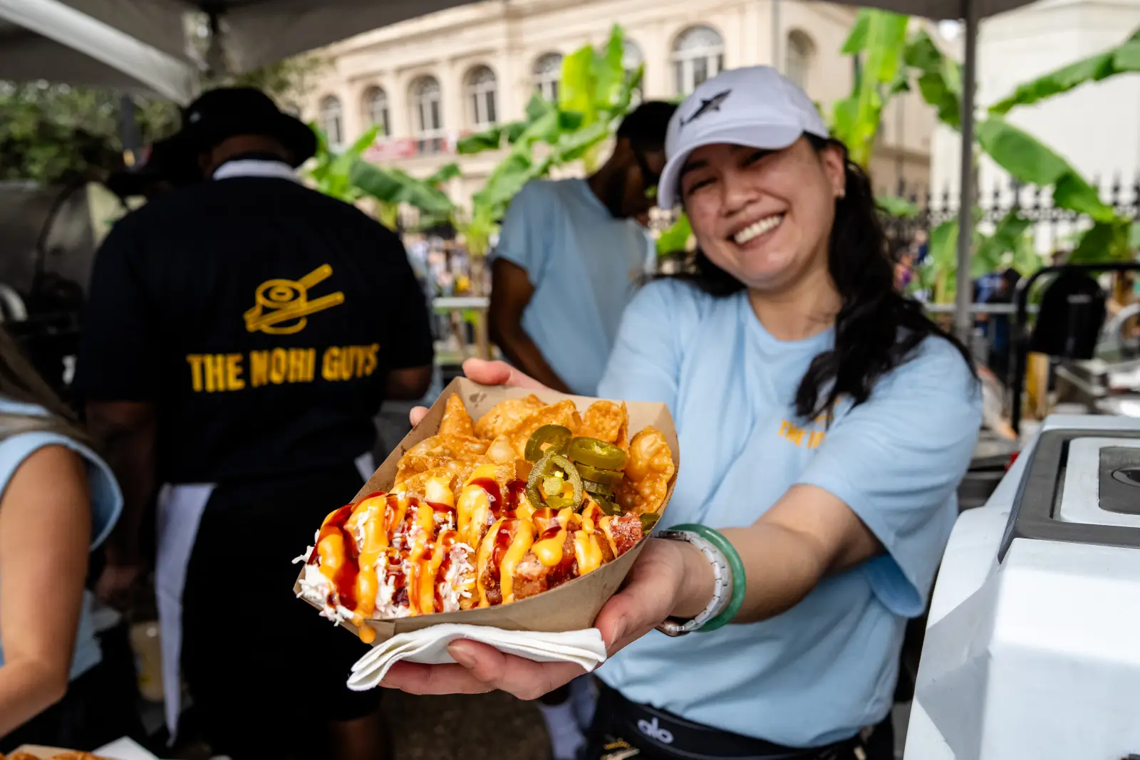 French Quarter Festival 2026 Draws Record Crowds, $4.3M In Vendor Earnings And 94% Hotel Occupancy Across New Orleans 3 A smiling vendor in a light blue Nori Guys shirt and white cap holds out a paper tray of sushi tacos topped with cheese sauce, sriracha drizzle, jalapeños and crispy chips at The Nori Guys stall during the 2026 French Quarter Festival, with a historic New Orleans building and tropical foliage visible in the background.