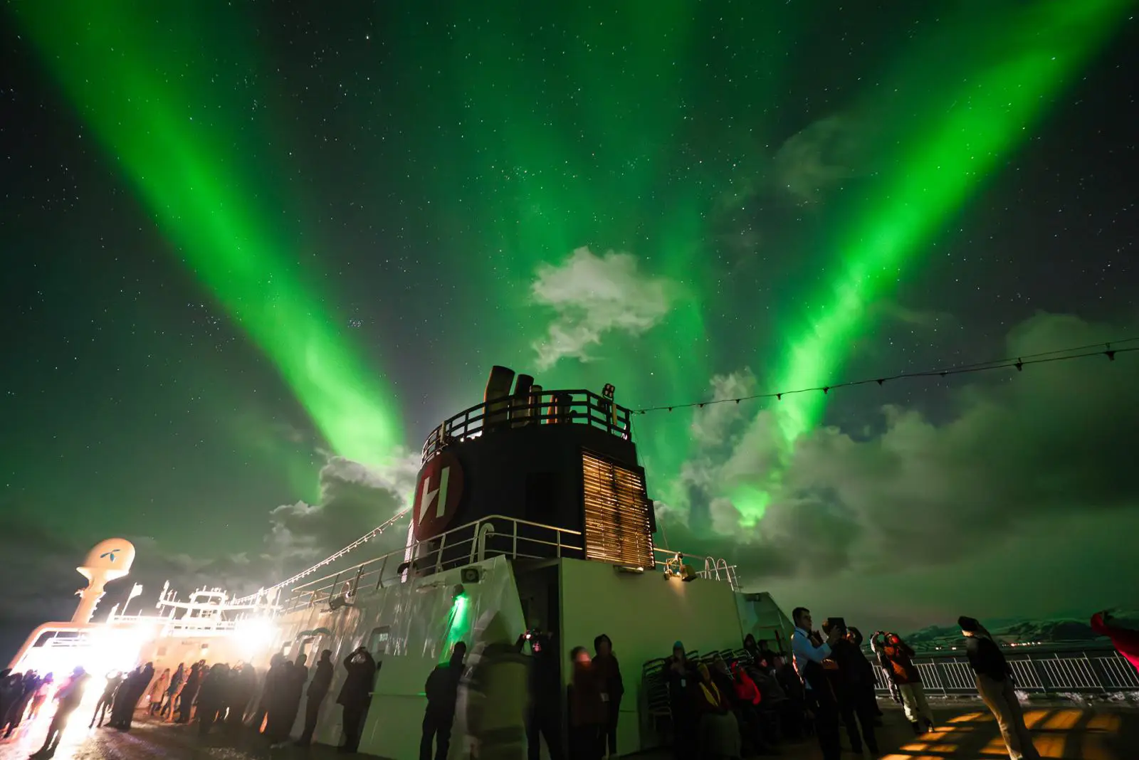 Passengers on the deck of a Hurtigruten ship watch bright green Northern Lights sweeping across the night sky in Arctic Norway.