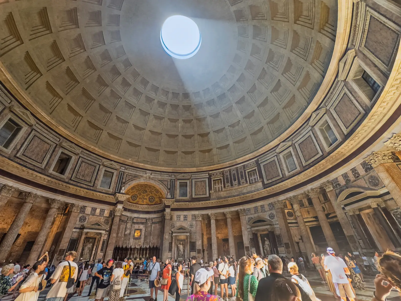 Wide-angle interior view of the Pantheon in Rome showing the coffered concrete dome and the circular oculus open to the sky at its apex, with a beam of natural light descending into the crowded rotunda and the ornate marble walls, columns and altar visible below