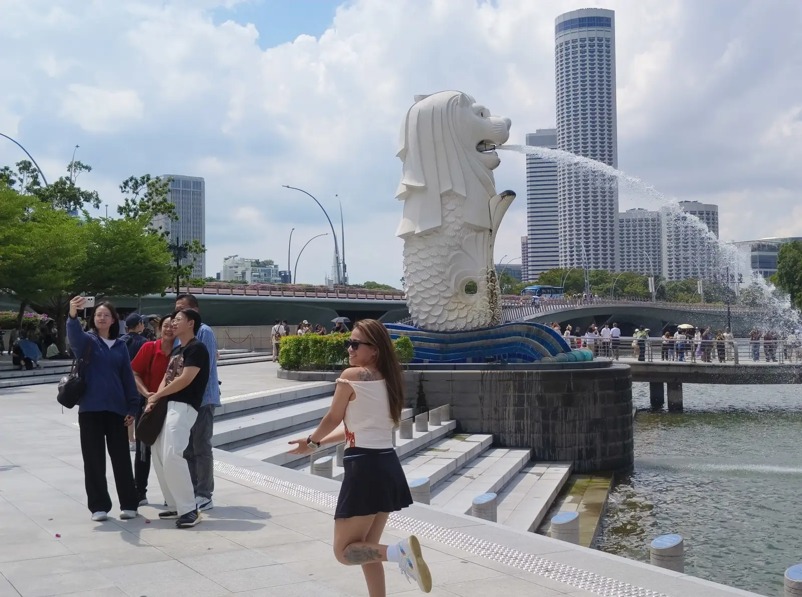 Tourists taking photos near the Merlion statue in Singapore with skyline and waterfront in the background on a sunny day