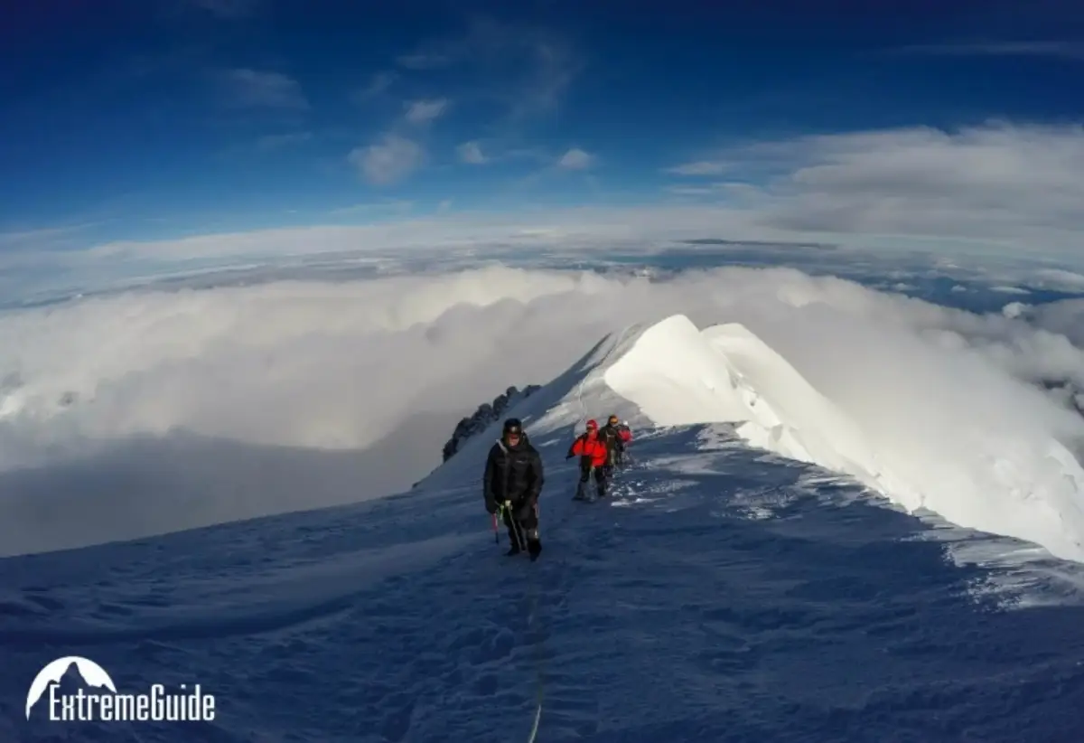 Climbers ascending a narrow snowy ridge above clouds during a high-altitude mountain expedition